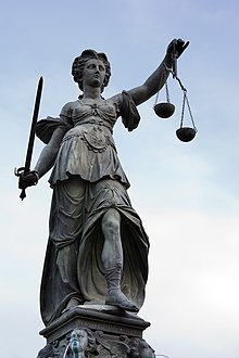 [1, 2] Die rechtsprechende Justitia wird mit Schwert und mit einer Waage dargestellt, mittels der sie mit verbundenen Augen (gerecht) das Für und Wider einer Entscheidung abwägt