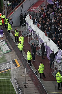 [3] Ordner (in neongelb) passen auf Fans der Gastmannschaft auf, dass die nicht über den Zaun klettern.