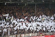 [1] Fans des FC St. Pauli, die bei Heimspielen in den Farben braun und weiß spielen, jubeln ihrer Mannschaft zu.